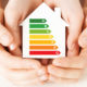 Ein Haus mit der Skala für die Energiewerte einer Immobilie wird von zwei Haenden festgehalten.